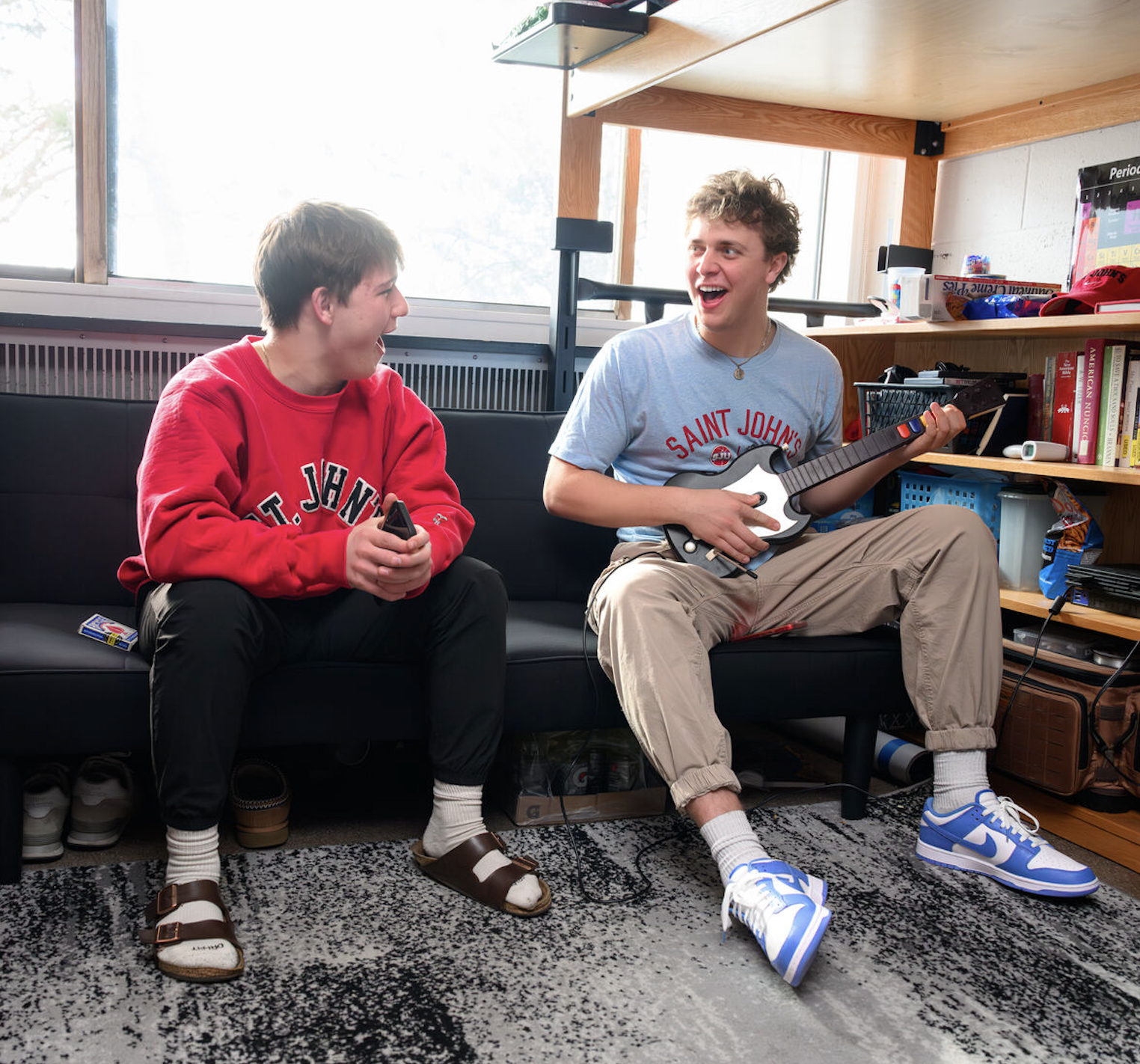 Two young men sit on a couch in a dorm room. One wears a red "St. John's" sweatshirt; the other, a gray "Saint John's" shirt, laughs while holding a video game guitar controller. The room has bookshelves and a window. They appear to be enjoying their time.